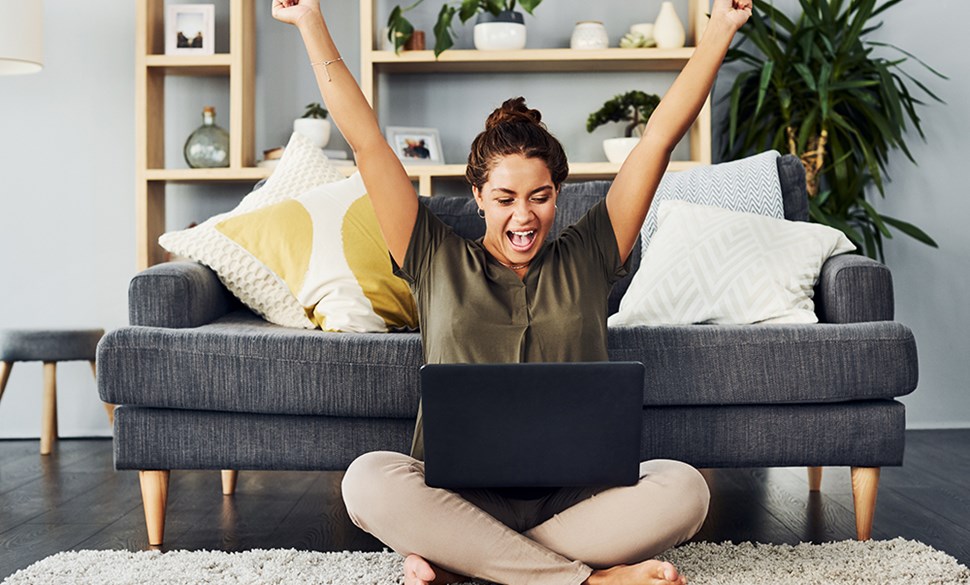 Kvinna sitter framför datorn och precis fått ett jobb, hon sträcker upp händerna av glädje.